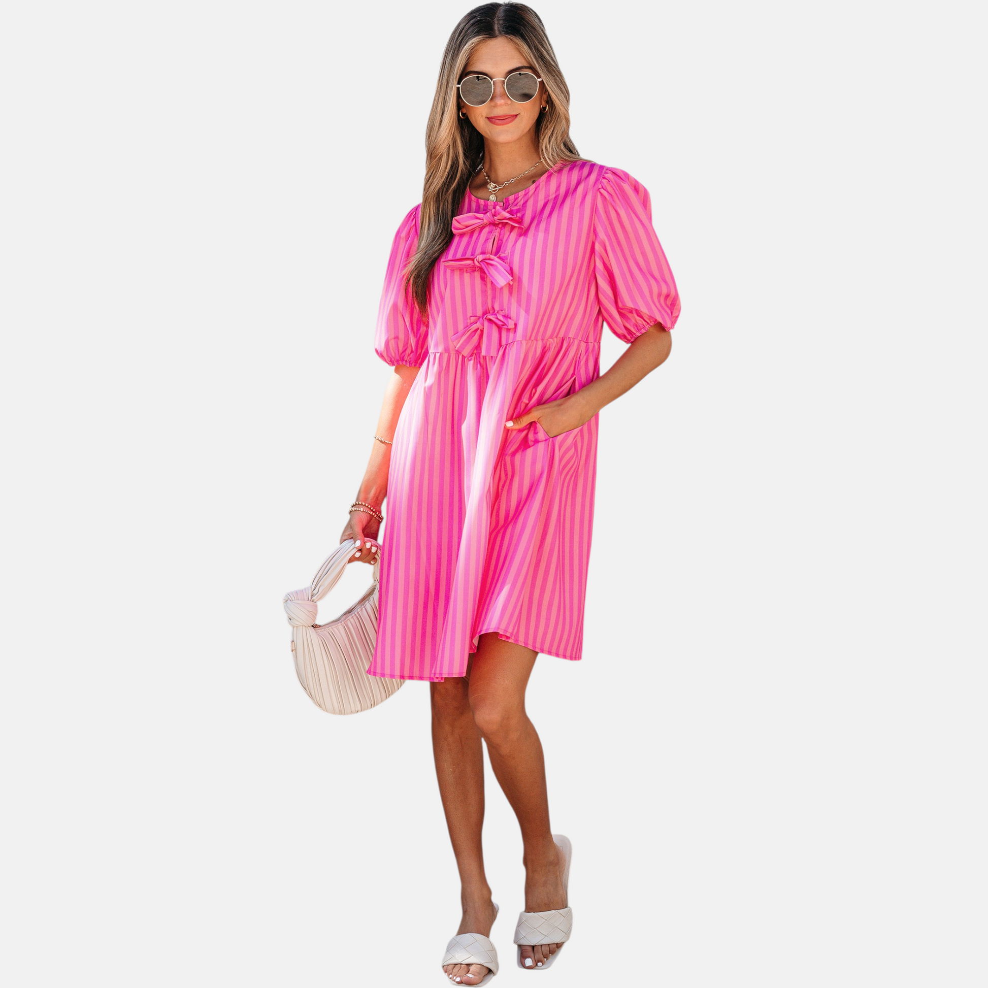A model posing in a vibrant pink striped mini dress featuring puff sleeves, three decorative front bows, and functional side pockets, styled with white slide sandals and a pleated handbag.