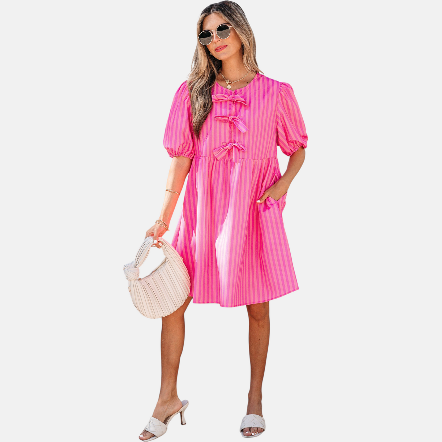 A model posing in a vibrant pink striped mini dress featuring puff sleeves, three decorative front bows, and functional side pockets, styled with white slide sandals and a pleated handbag.