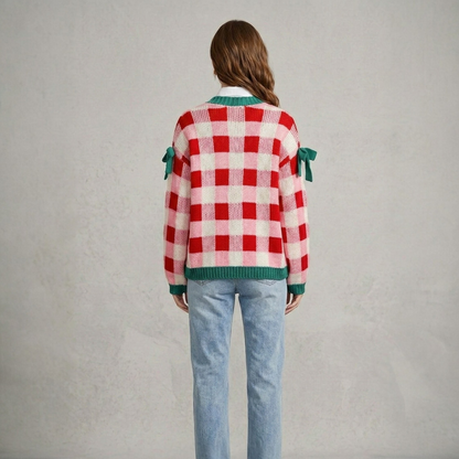 Back view of a model wearing a pink and red checkered open-front knit cardigan with green trim and decorative sleeve bows, styled with a white shirt and blue jeans.