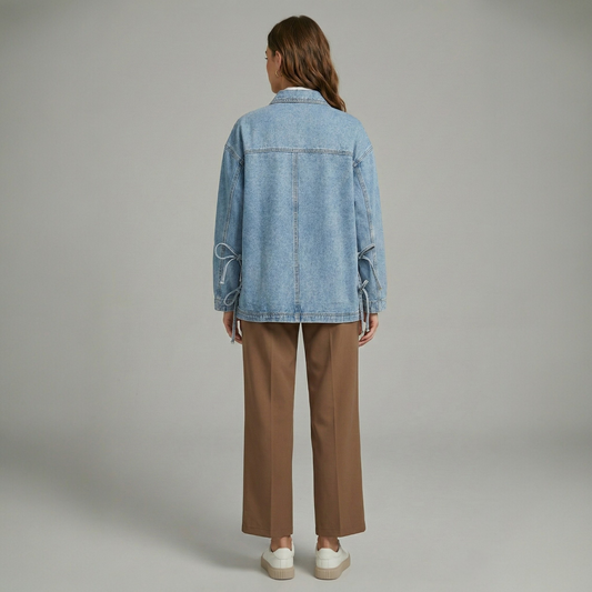 Back view of a model wearing a light-wash denim boyfriend jacket with unique side tie-up strings, paired with brown trousers.