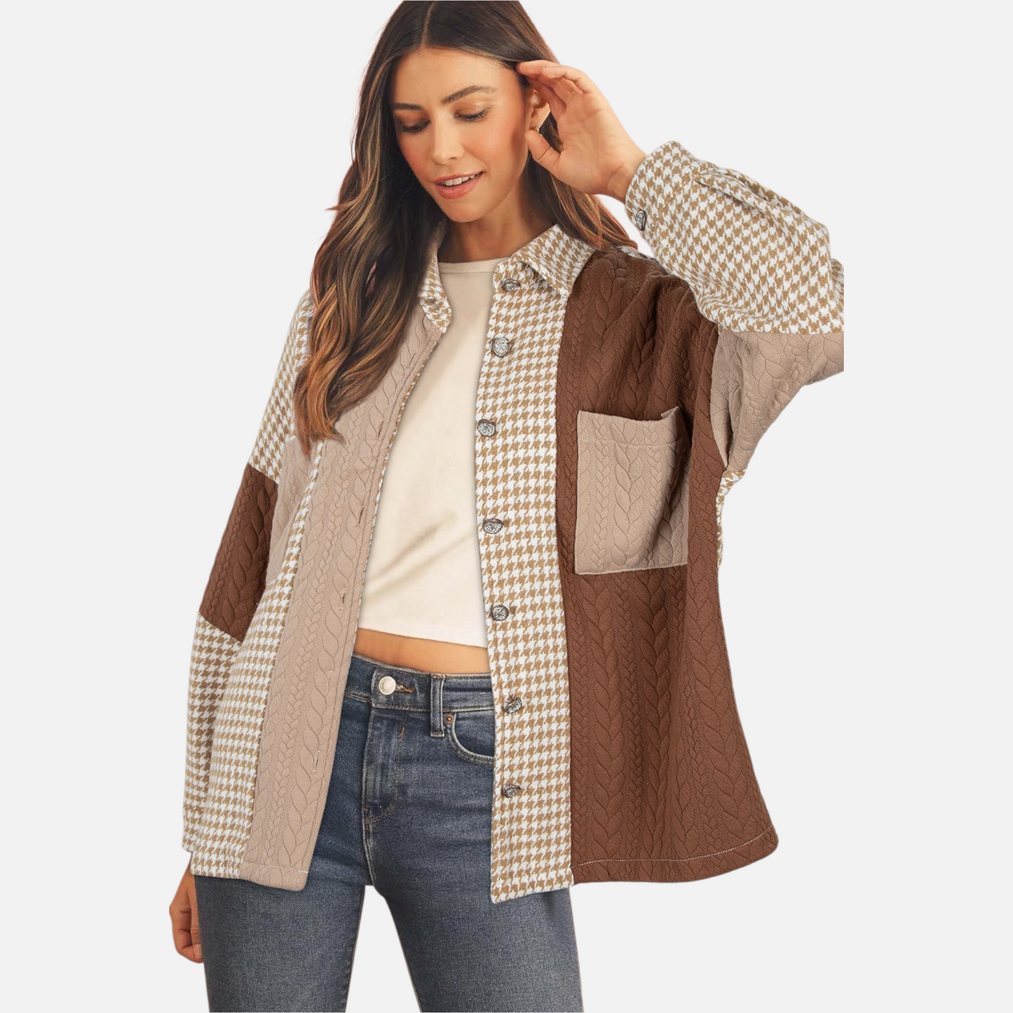 A model styled in a brown patchwork shacket with houndstooth and cable-knit textures, layered over a white top and blue jeans, looking down with a smile.