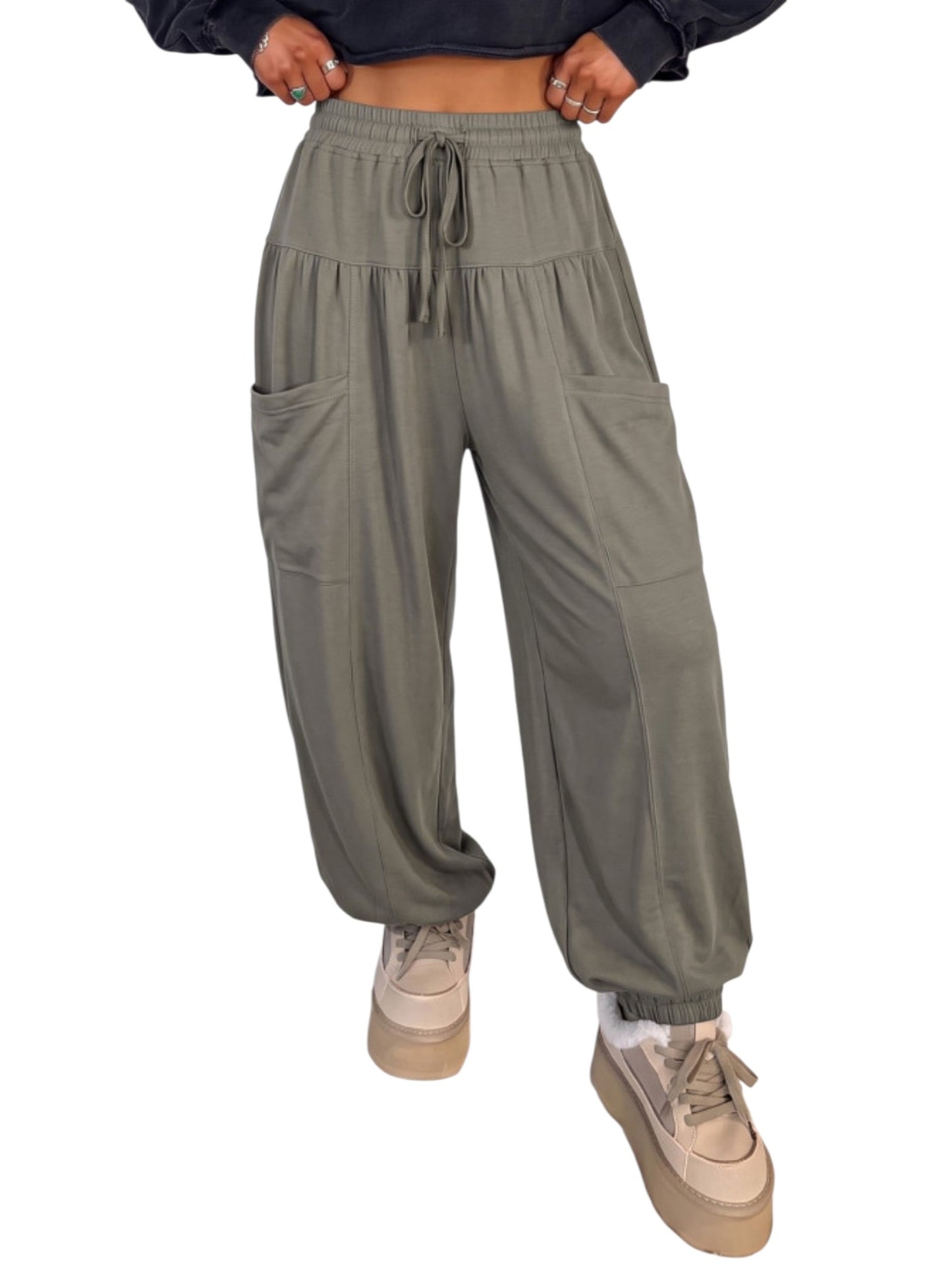 A person wearing loose, wide-leg jogger-style pants in a muted sage green color, paired with beige platform sneakers and a dark cropped top. The photo is cropped from the chest down on a plain white background.