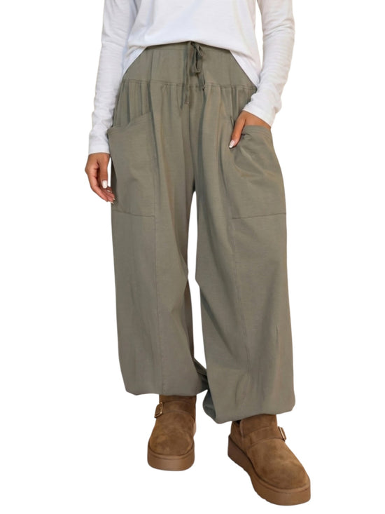 A person wearing loose, wide-leg olive green drawstring pants with large front patch pockets, paired with a white long-sleeve top and brown suede boots, shown against a plain white background.