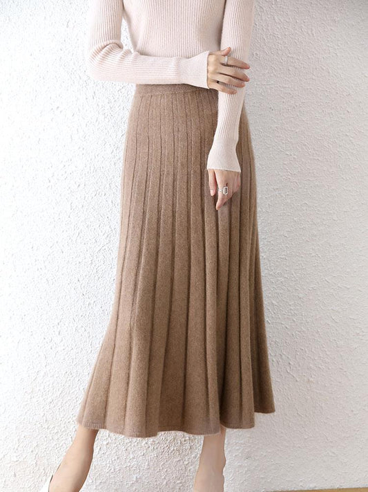 Person wearing a beige sweater and brown pleated skirt against a white background