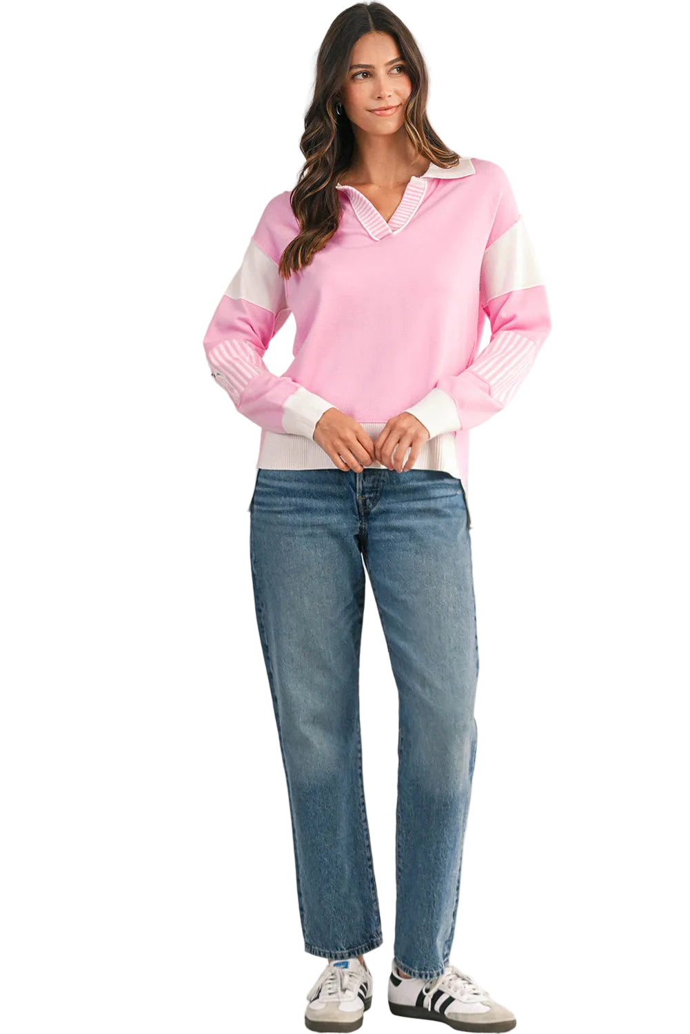 Woman wearing a pink sweater with white accents and blue jeans, standing against a light background. 