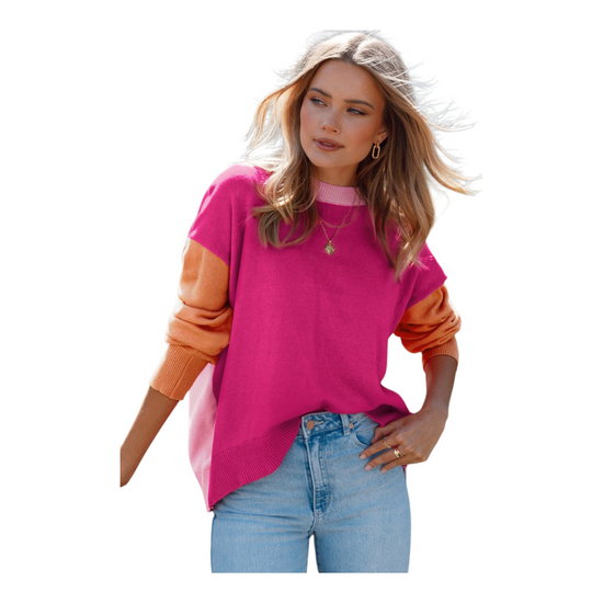 A woman wearing an oversized knit sweater with a bold color-block design. The front of the sweater is a vibrant magenta with a light pink crew neckline. She is paired with light-wash denim jeans and gold jewelry.