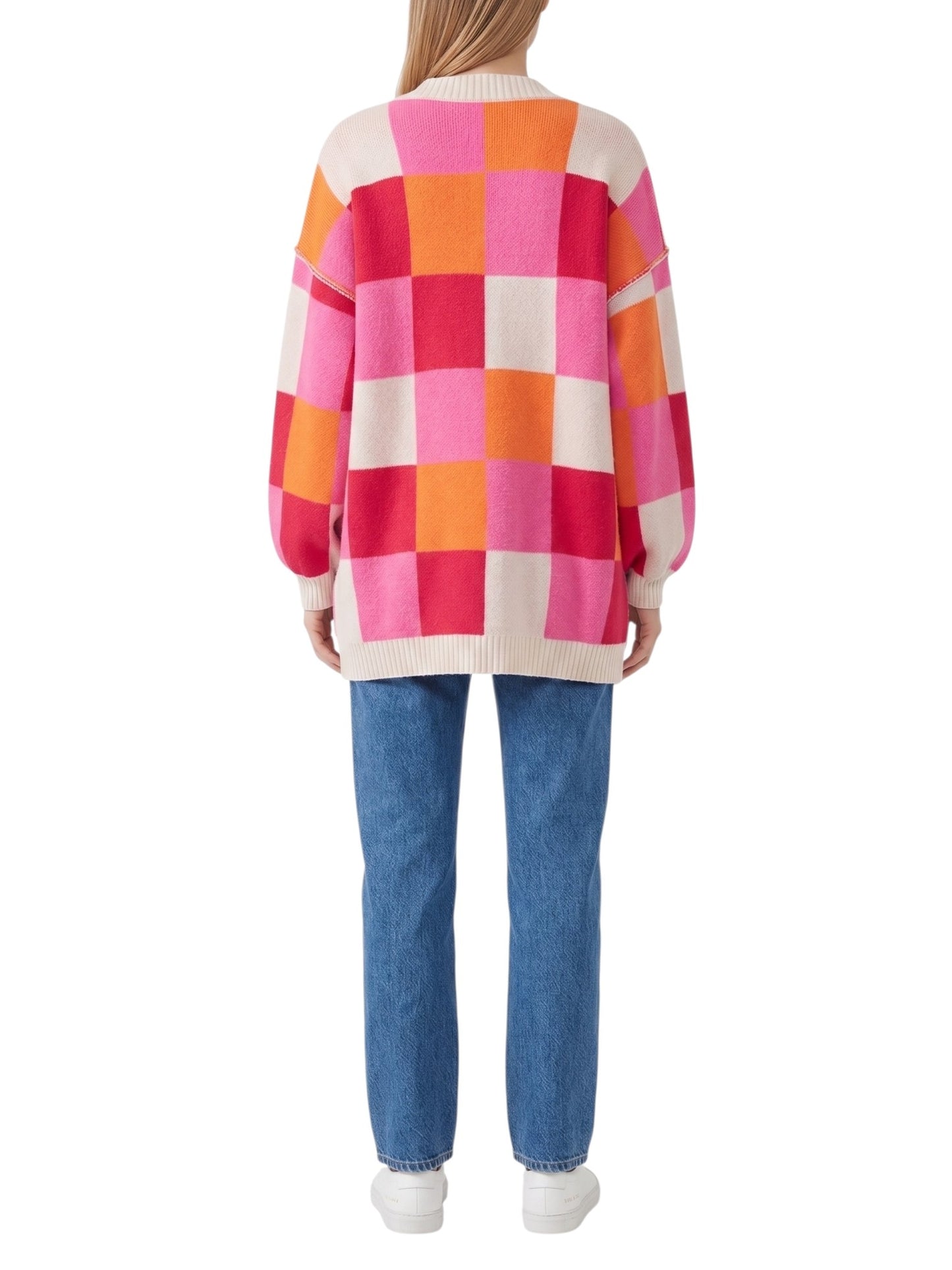 A woman viewed from the back wearing a colorful oversized knit sweater with a pink, orange, and cream checkered pattern, paired with blue jeans and white sneakers, on a plain white background.
