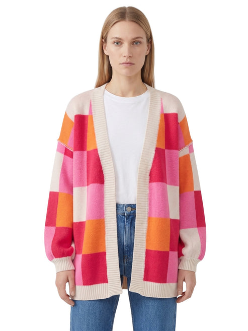 A woman modeling a chunky knit cardigan with a large checkered pattern in pink, orange, and cream, worn over a white t-shirt and blue jeans against a plain white background.