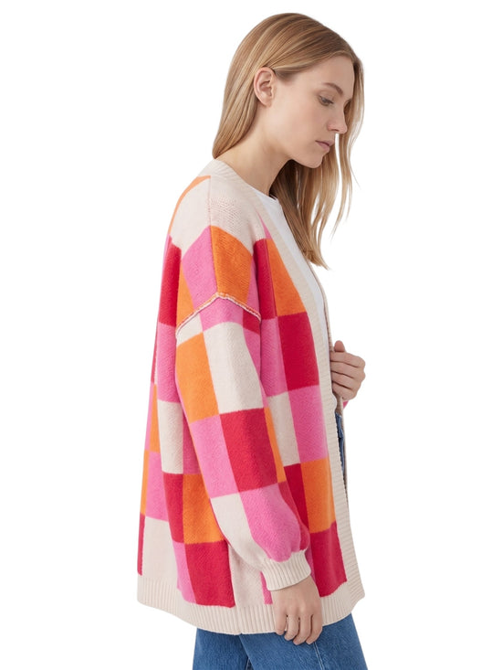 A woman wearing a chunky knit cardigan with a large checkered pattern in pink, orange, and cream, paired with blue jeans, shown from a side profile against a white background.