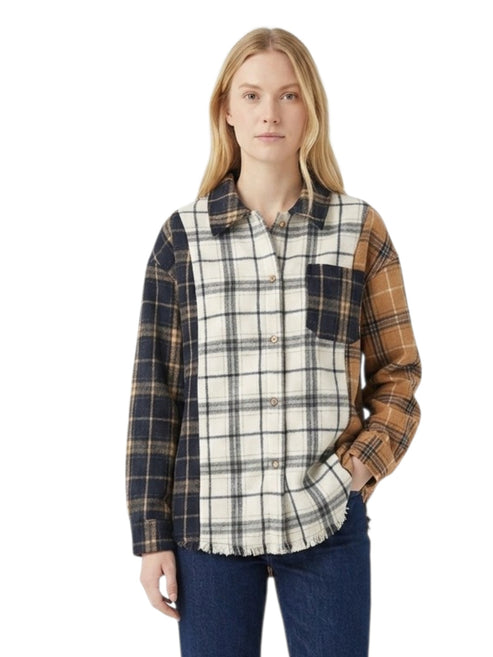 A woman modeling a long-sleeve patchwork plaid button-up shirt with navy, tan, and cream sections, paired with blue jeans against a plain white background.