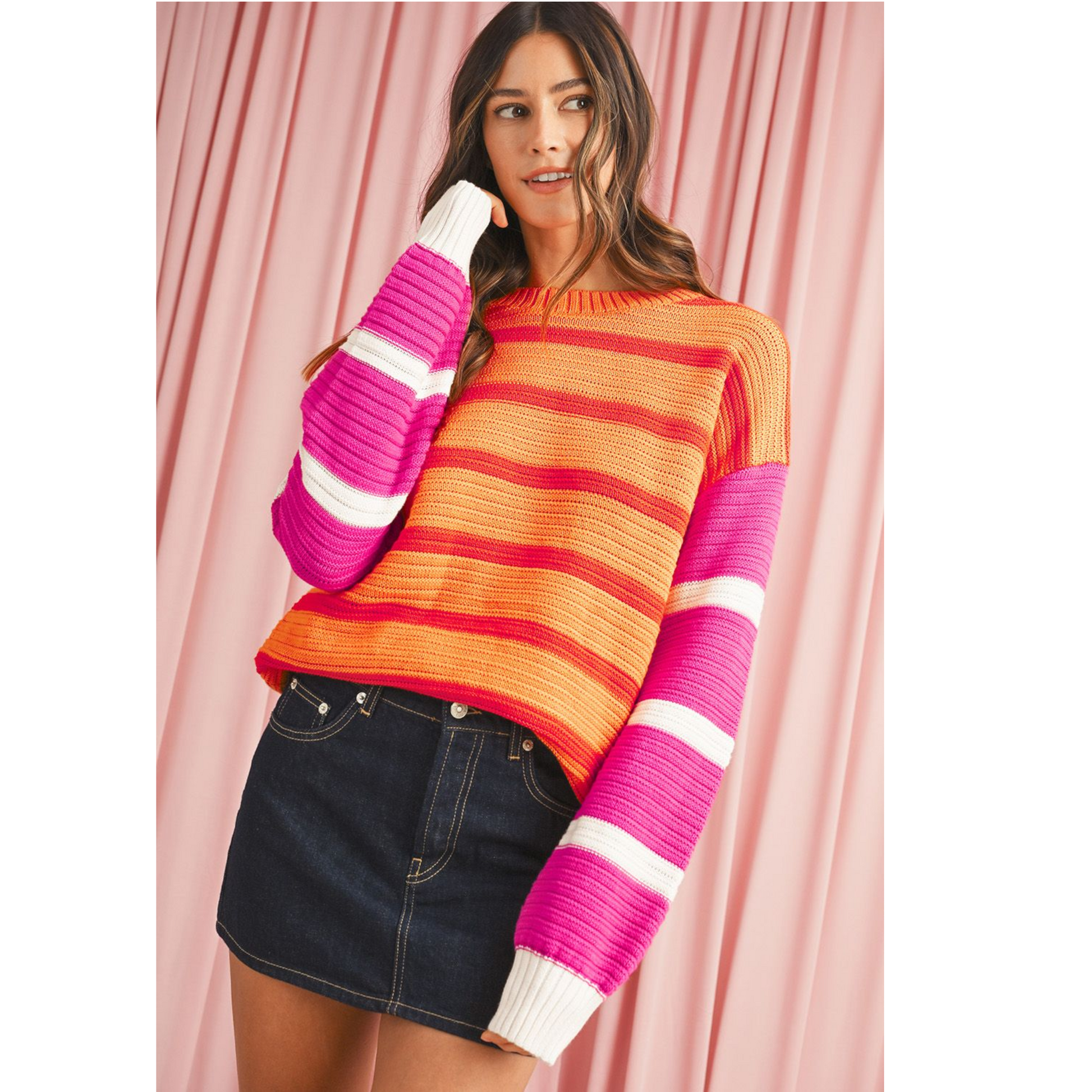 Woman wearing a colorful striped sweater and denim skirt against a pink curtain.