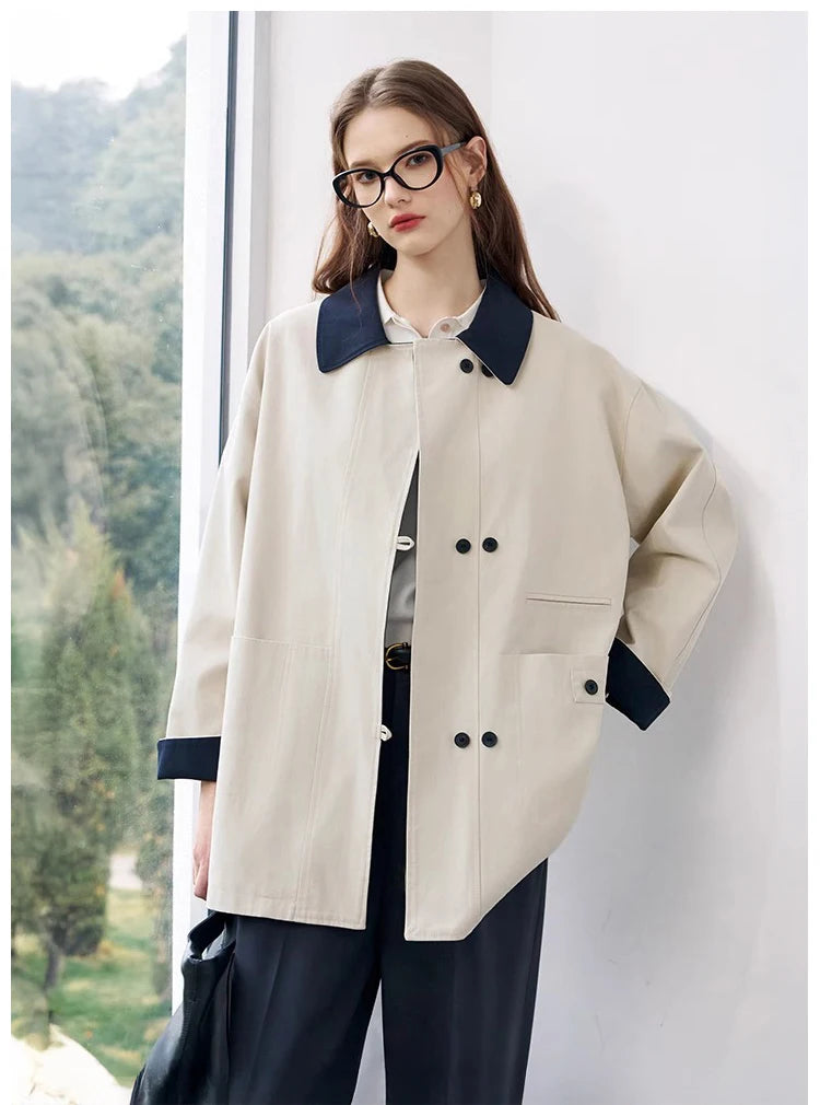 Woman wearing a beige coat with a dark collar and buttons, standing against a white wall with a blurred natural background.