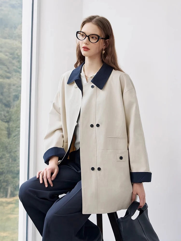 Woman wearing a beige coat with navy trim sitting indoors.