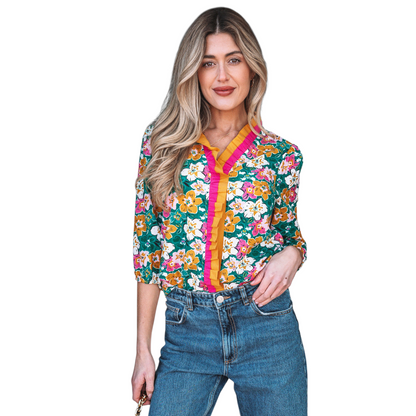 A woman wearing an emerald green floral V-neck blouse featuring distinctive magenta and ochre contrast frilled trim along the neckline and 3/4 length sleeves.