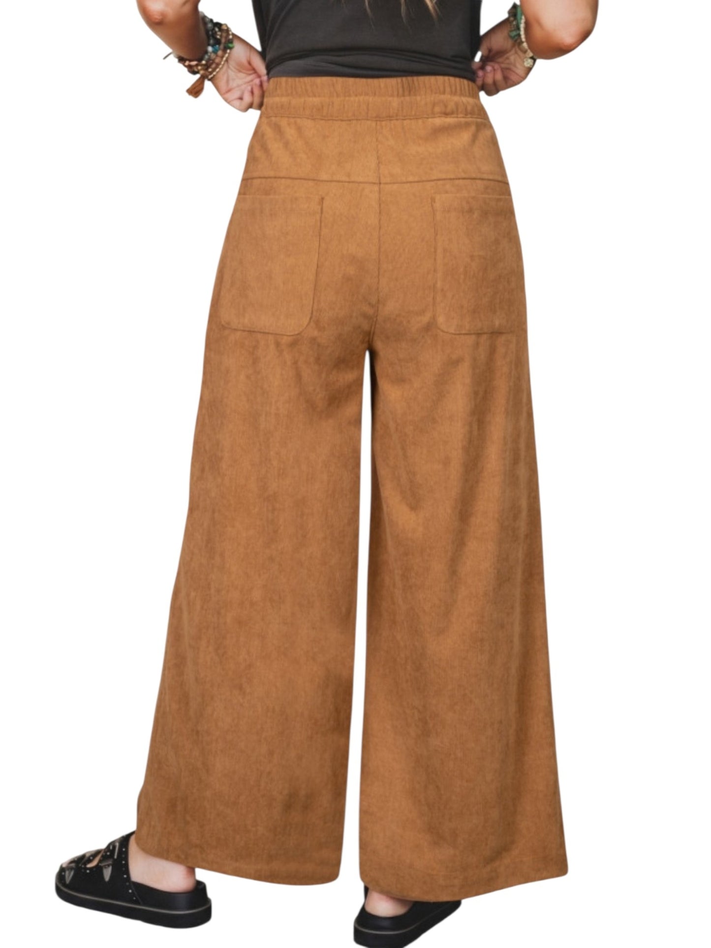 A person shown from the back wearing high-waisted, wide-leg brown pants with a textured corduroy-like finish, a black top, and black slide sandals against a plain white background.