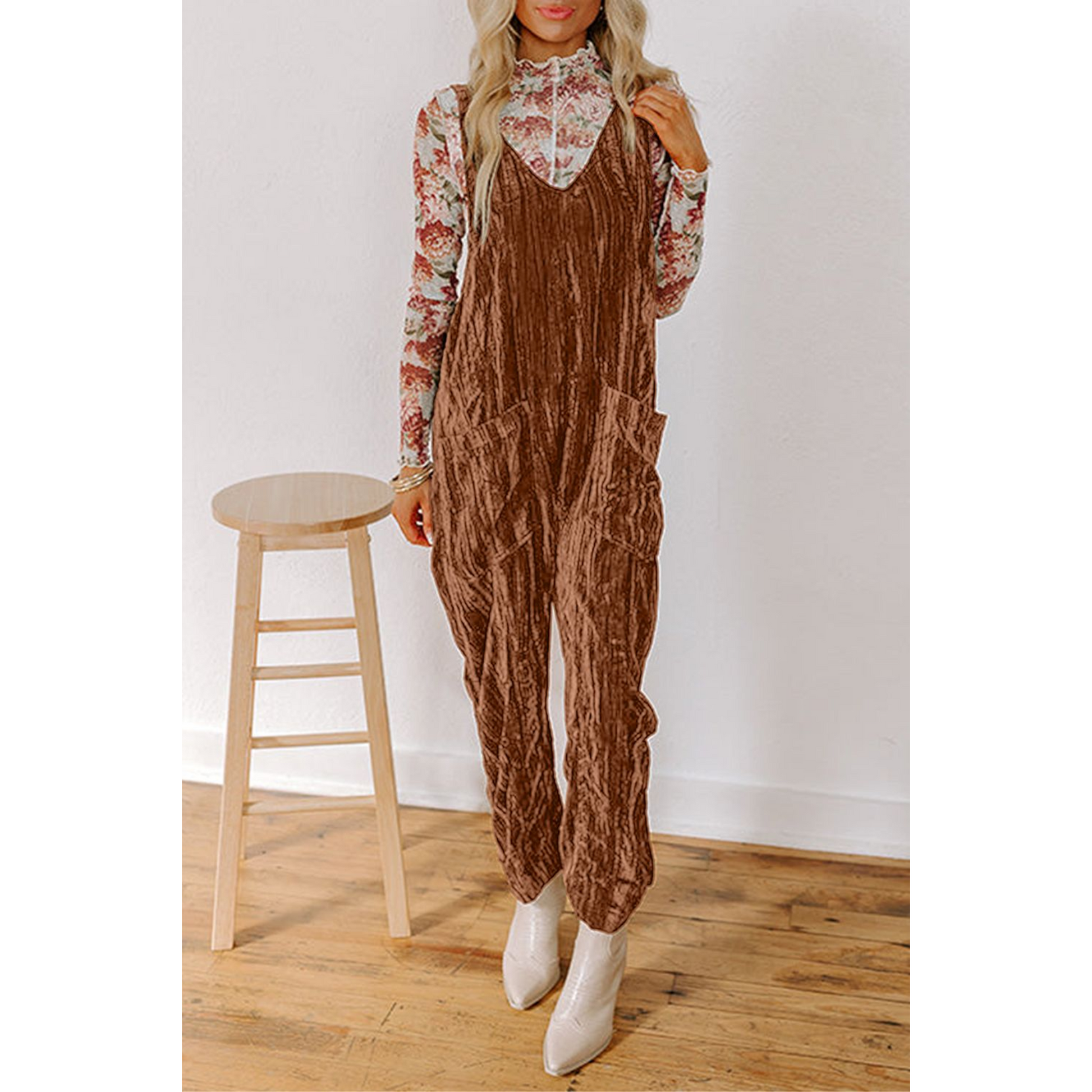 Woman wearing a brown textured jumpsuit with a floral top, standing next to a wooden stool against a white wall.