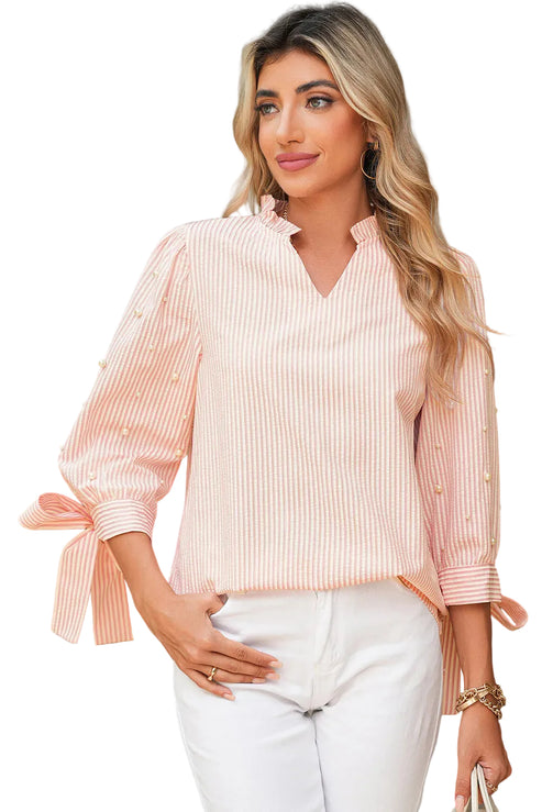 Woman wearing a pink striped blouse with a tie detail and white pants, standing outdoors.