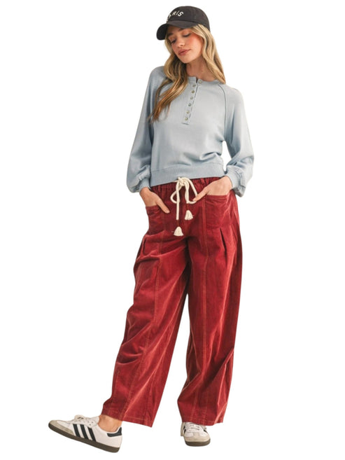 A woman modeling a light blue long-sleeve henley top and red corduroy wide-leg pants with a cream drawstring, paired with a black baseball cap and white sneakers, on a solid white background.
