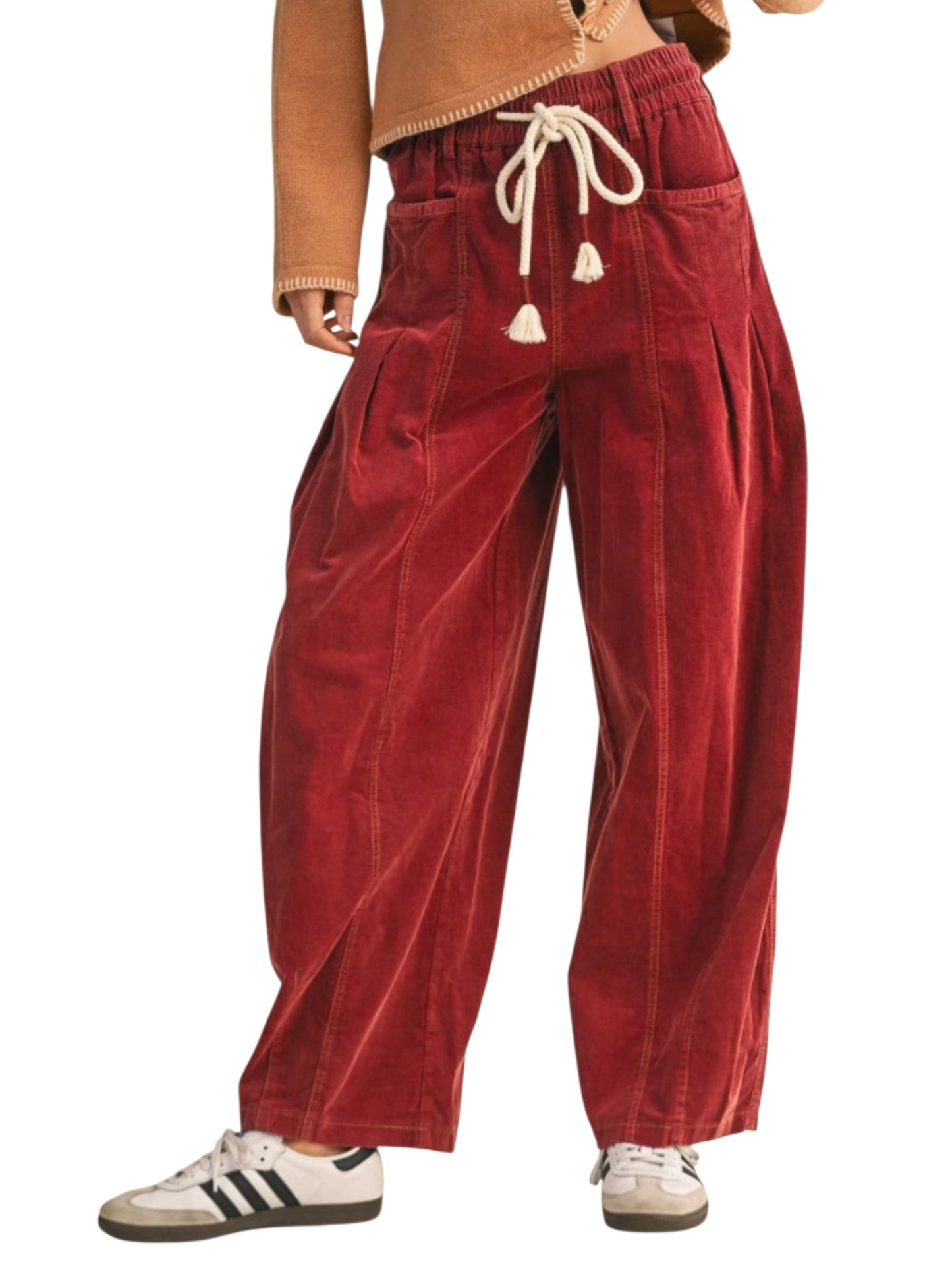 A person wearing loose, wide-leg corduroy pants in a deep red or burgundy shade, paired with white sneakers and a tan top, shown against a plain white background.