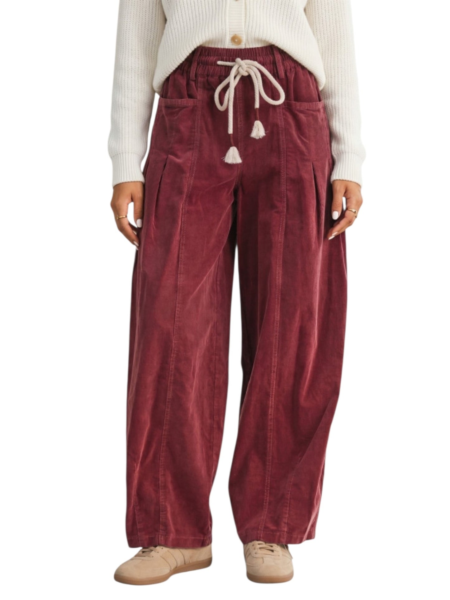 A person wearing loose burgundy corduroy drawstring pants with side pockets, paired with a cream ribbed cardigan and beige shoes, shown against a plain white background.