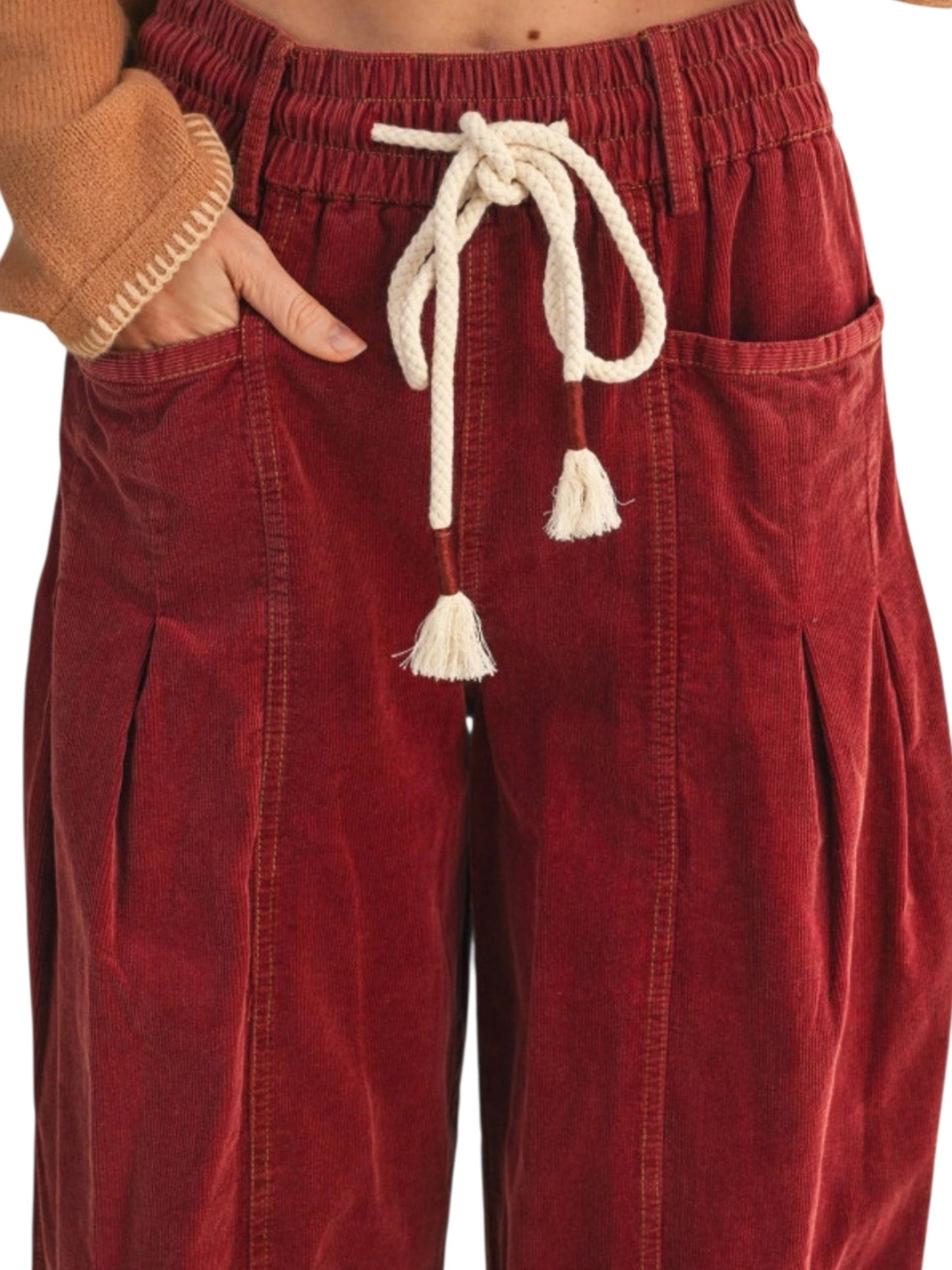 A close-up of a person wearing wide-leg corduroy pants in a deep red or burgundy color, featuring an elastic waistband, cream-colored drawstring with tassels, and side pockets.