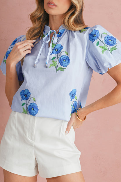 Woman wearing a light blue blouse with floral embroidery and white shorts against a pink background