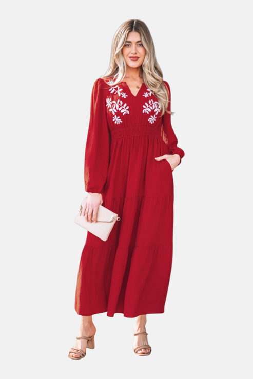 A full-length front view of a model wearing a burgundy tiered maxi dress featuring intricate white floral embroidery on the bodice and a flattering cinched waist.