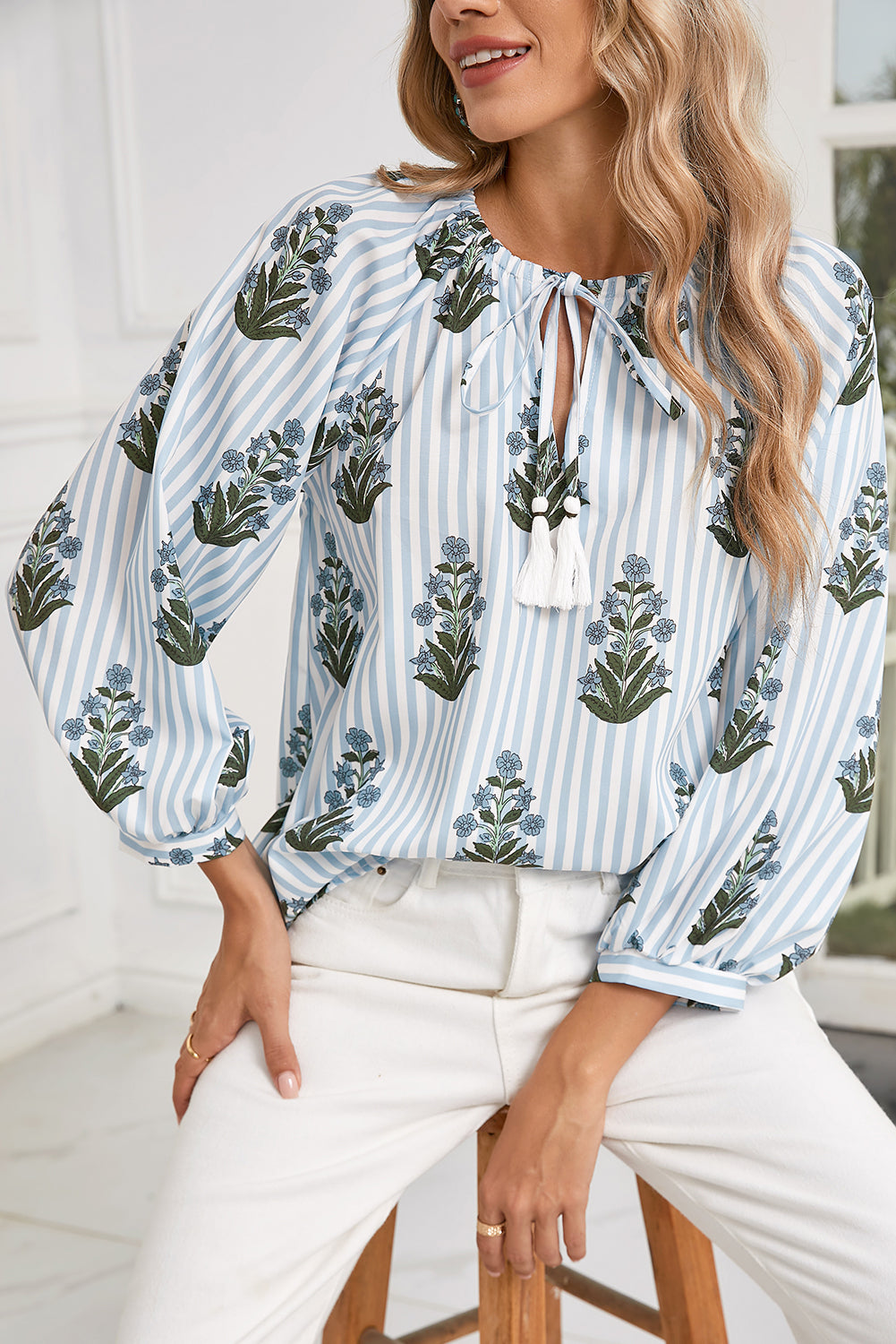 Front view of a blue floral striped blouse with long sleeves, perfect for spring and summer, worn by a model sitting on a wooden stool in a boho-chic style.