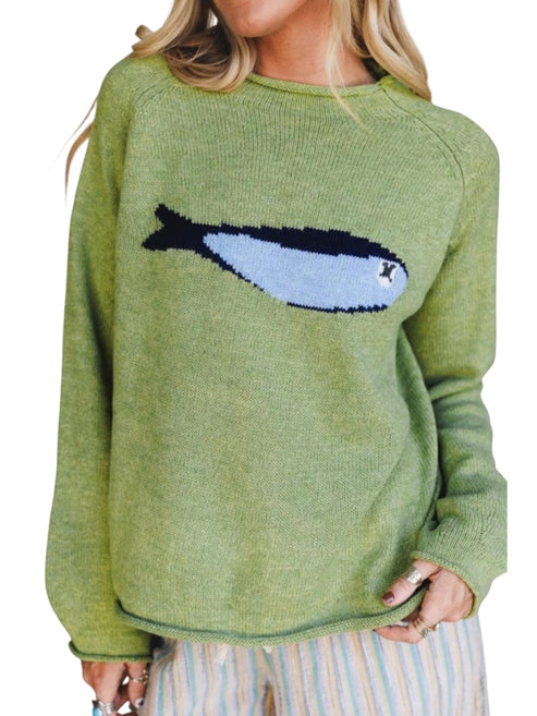 A woman wearing a sage green knit sweater with a large knitted fish graphic across the chest in light blue and black, paired with striped pants against a white background.