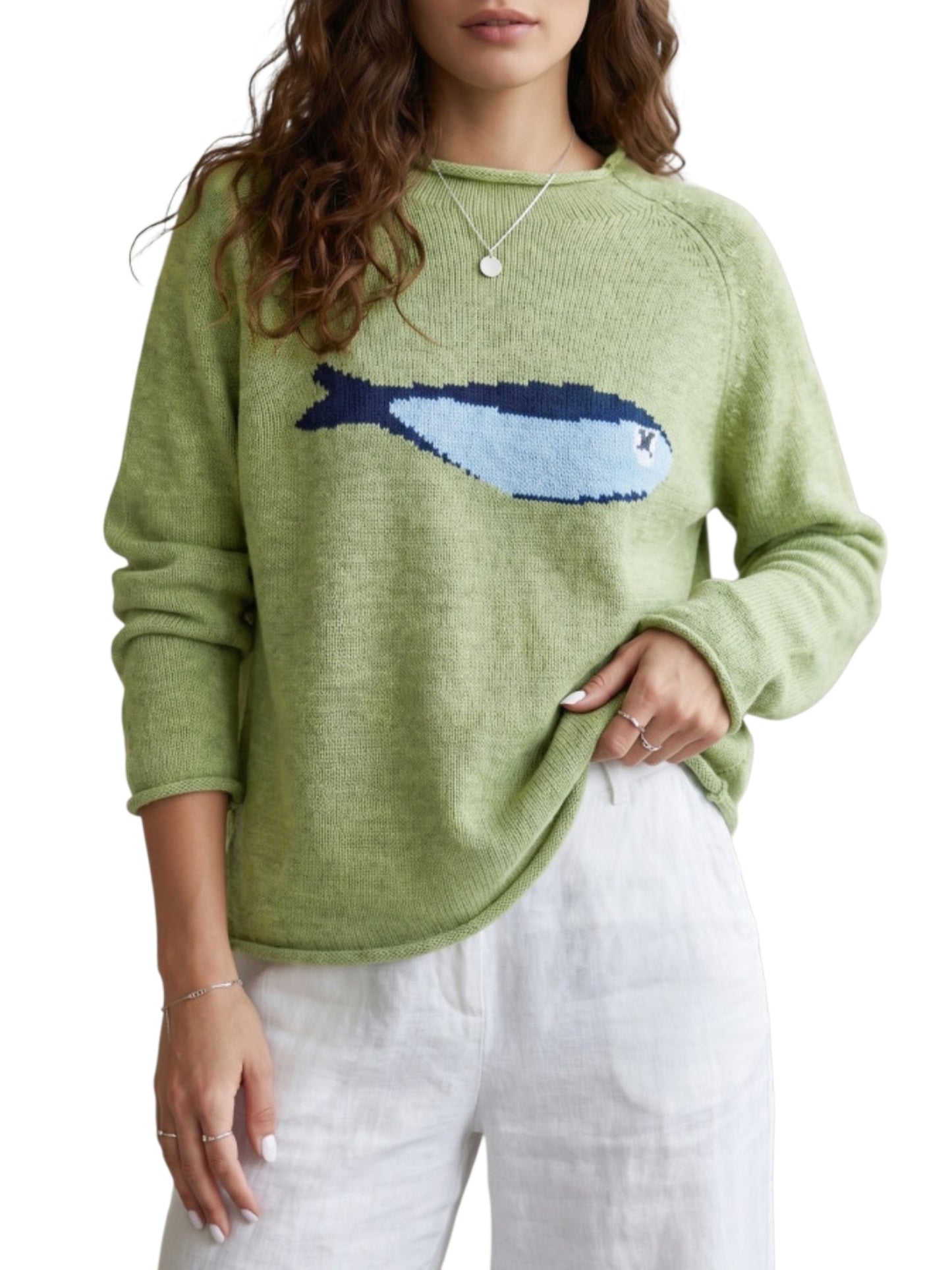 A woman wearing a light green knit sweater with a large blue and white fish motif across the chest, paired with white pants, against a plain white background.