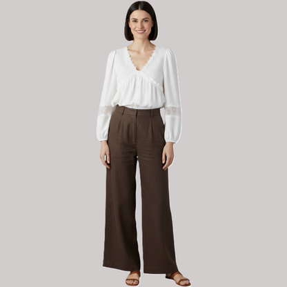 A full-length studio shot of a woman wearing a white floral guipure lace V-neck blouse tucked into dark brown wide-leg trousers, styled with flat brown sandals.