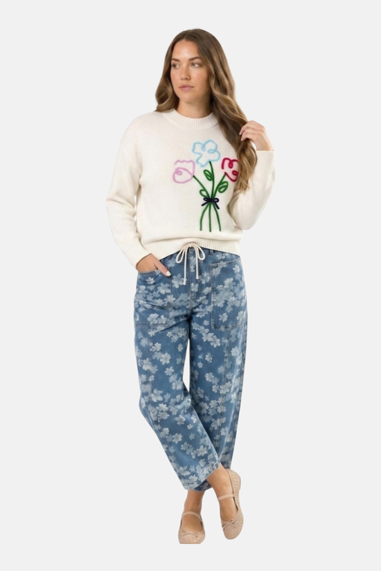 A model showcasing a transitional spring outfit featuring blue floral barrel-leg jeans paired with a cream crew-neck knit jumper with embroidered flower details.