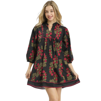 A woman wearing a black 100% cotton mini dress featuring a red floral print, a structured squared yoke with contrast piping, and a notched v-neckline.