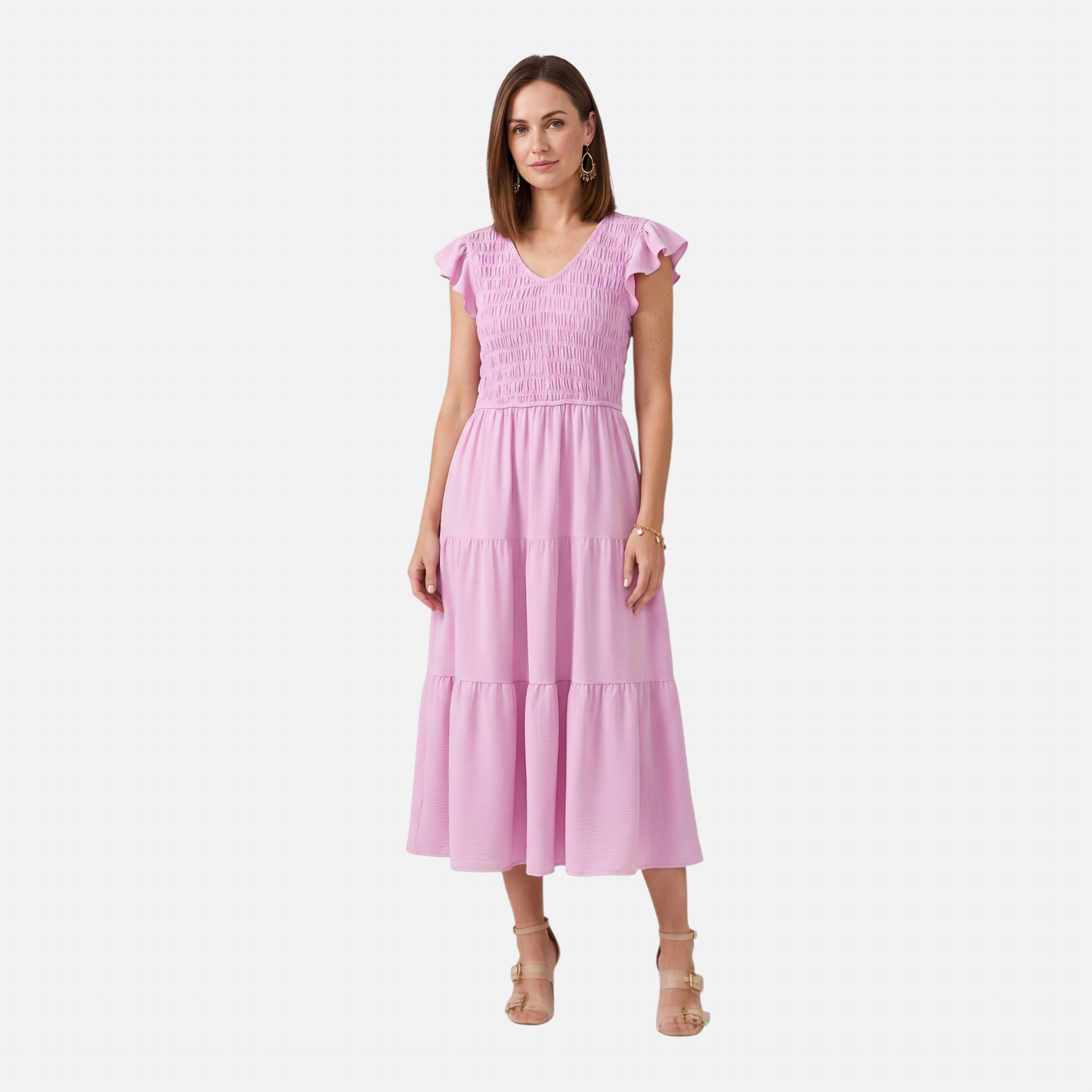 A woman wearing a light pink midi dress with a V-neckline and a fully smocked bodice. The dress features short flutter sleeves and a tiered, flowing skirt. She is paired with tan block-heel sandals and gold drop earrings against a plain white background.