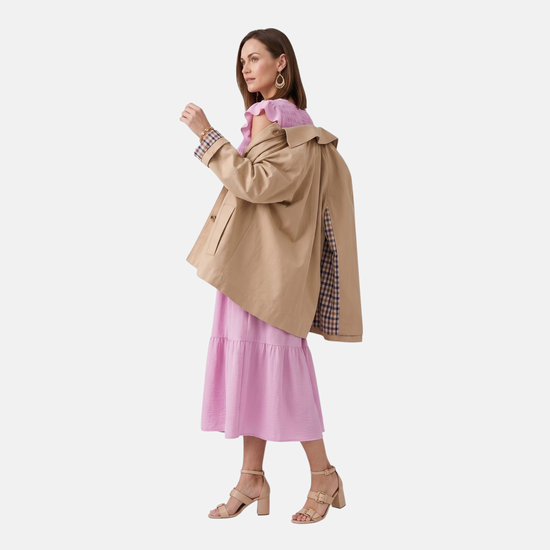 A woman wearing the pink smocked midi dress styled with a tan trench coat draped over her shoulders. The coat features a checkered lining on the cuffs and interior. She is shown from a side profile, highlighting the movement of the tiered skirt.