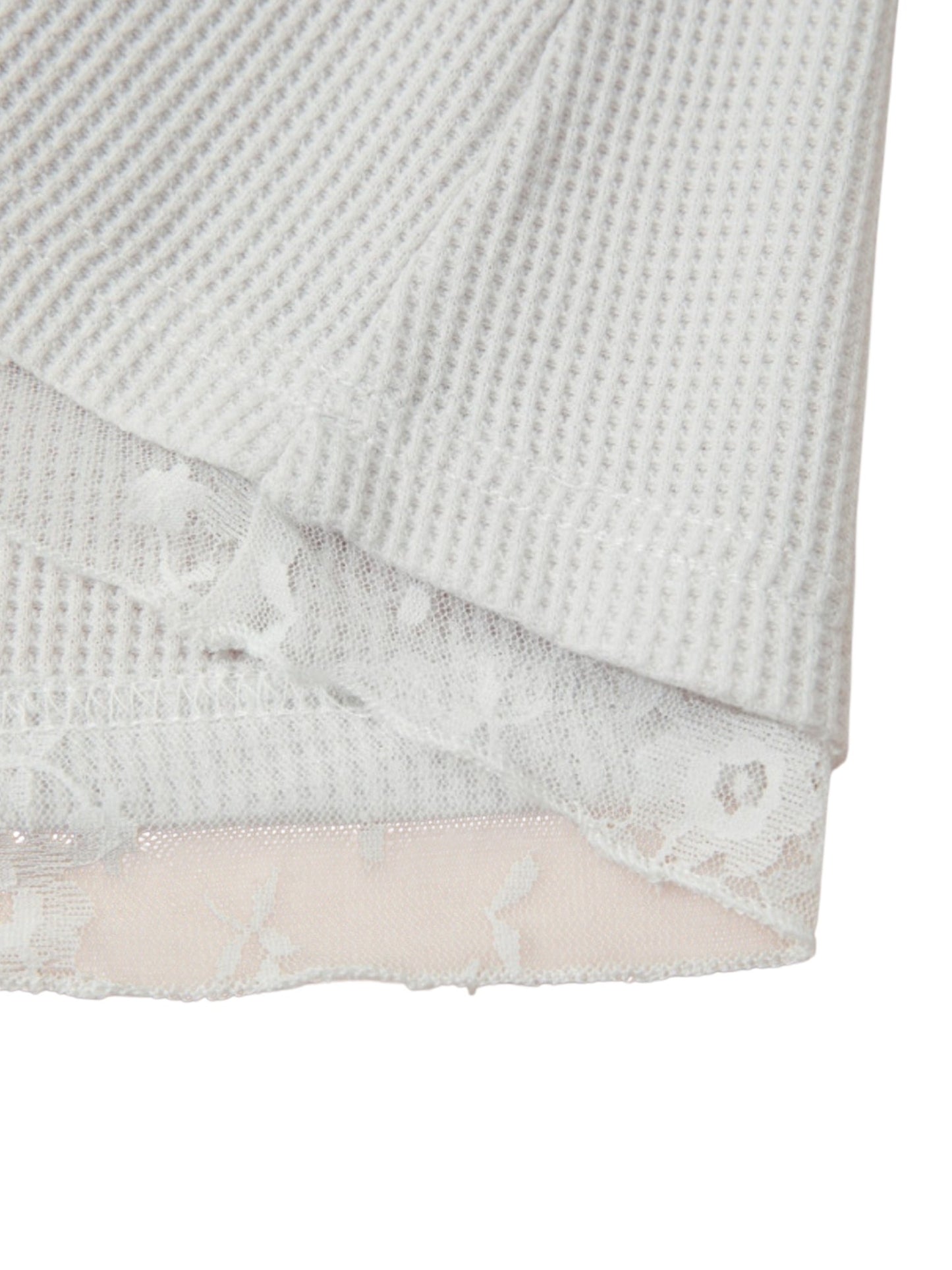 Close-up of a white textured waffle-knit fabric layered over a sheer white lace trim, with a pale pink lining visible along the edge.