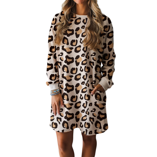 A woman wearing a cream and brown leopard print shift dress with long sleeves, a round neckline, and functional side pockets in a relaxed lifestyle setting.
