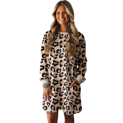 A woman wearing a cream and brown leopard print shift dress with long sleeves, a round neckline, and functional side pockets in a relaxed lifestyle setting.