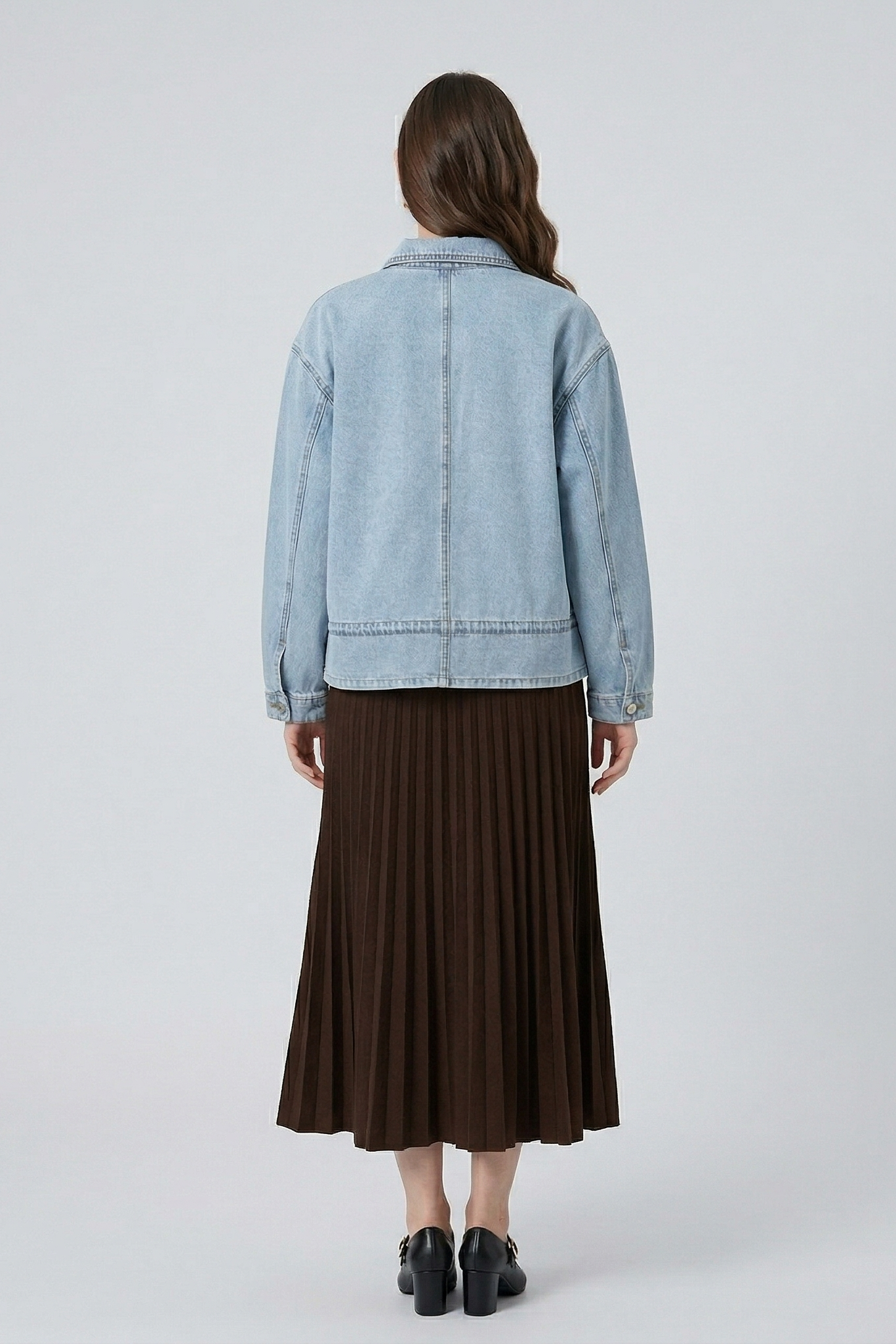 Rear view of a model wearing the light blue drawstring denim jacket styled with a dark brown pleated maxi skirt for a feminine, casual look.