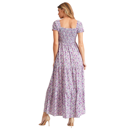 Model wearing a lilac and purple floral smocked maxi dress with short puff sleeves and a tiered skirt. The dress features a square neckline and an elasticated smocked bodice for a flattering fit. Back view.