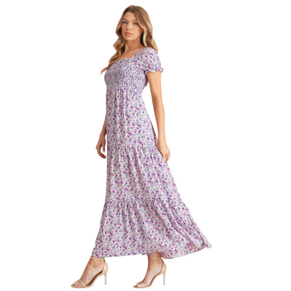 Model wearing a lilac and purple floral smocked maxi dress with short puff sleeves and a tiered skirt. The dress features a square neckline and an elasticated smocked bodice for a flattering fit. Side view.