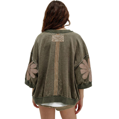 Rear view of an olive green mineral wash sweatshirt featuring a vertical central seam, 3/4 sleeves with floral patchwork, and a relaxed, oversized fit.