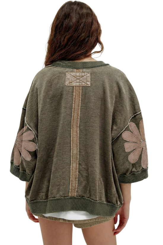 Rear view of an olive green mineral wash sweatshirt featuring a vertical central seam, 3/4 sleeves with floral patchwork, and a relaxed, oversized fit.