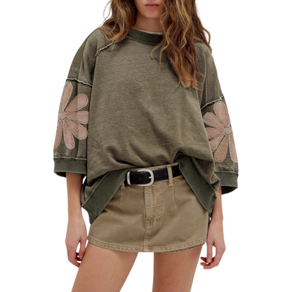 Close-up of a deconstructed sweatshirt in a mineral wash olive hue, highlighting the raw-edge diagonal seams and large mauve floral appliqués on the sleeves.