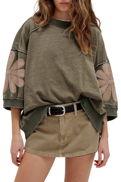 Close-up of a deconstructed sweatshirt in a mineral wash olive hue, highlighting the raw-edge diagonal seams and large mauve floral appliqués on the sleeves.
