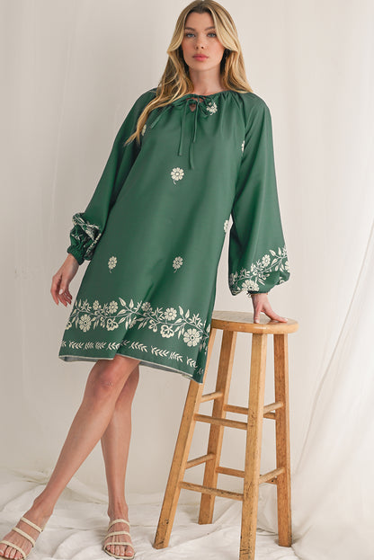 Woman in a mist green boho mini dress with long puff sleeves, leaning on a wooden chair for a chic, relaxed vibe.