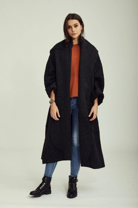 Woman wearing a black coat, orange sweater, blue jeans, and black boots on a light gray background