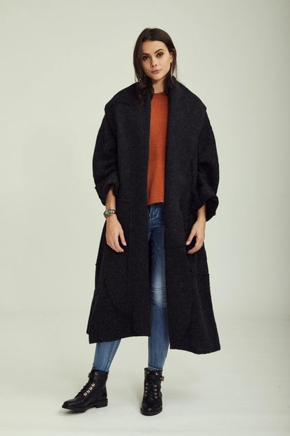 Woman wearing a black coat, orange sweater, blue jeans, and black boots on a light gray background