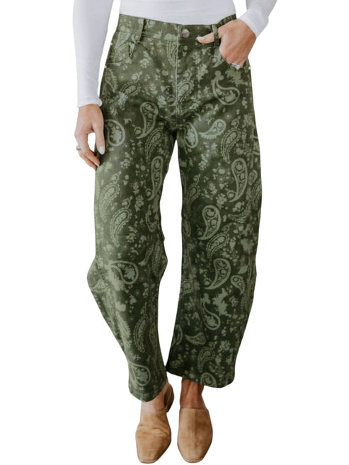 A person wearing loose, wide-leg pants with an olive green and light sage paisley pattern, styled with a white long-sleeve top and tan slip-on shoes against a plain white background.