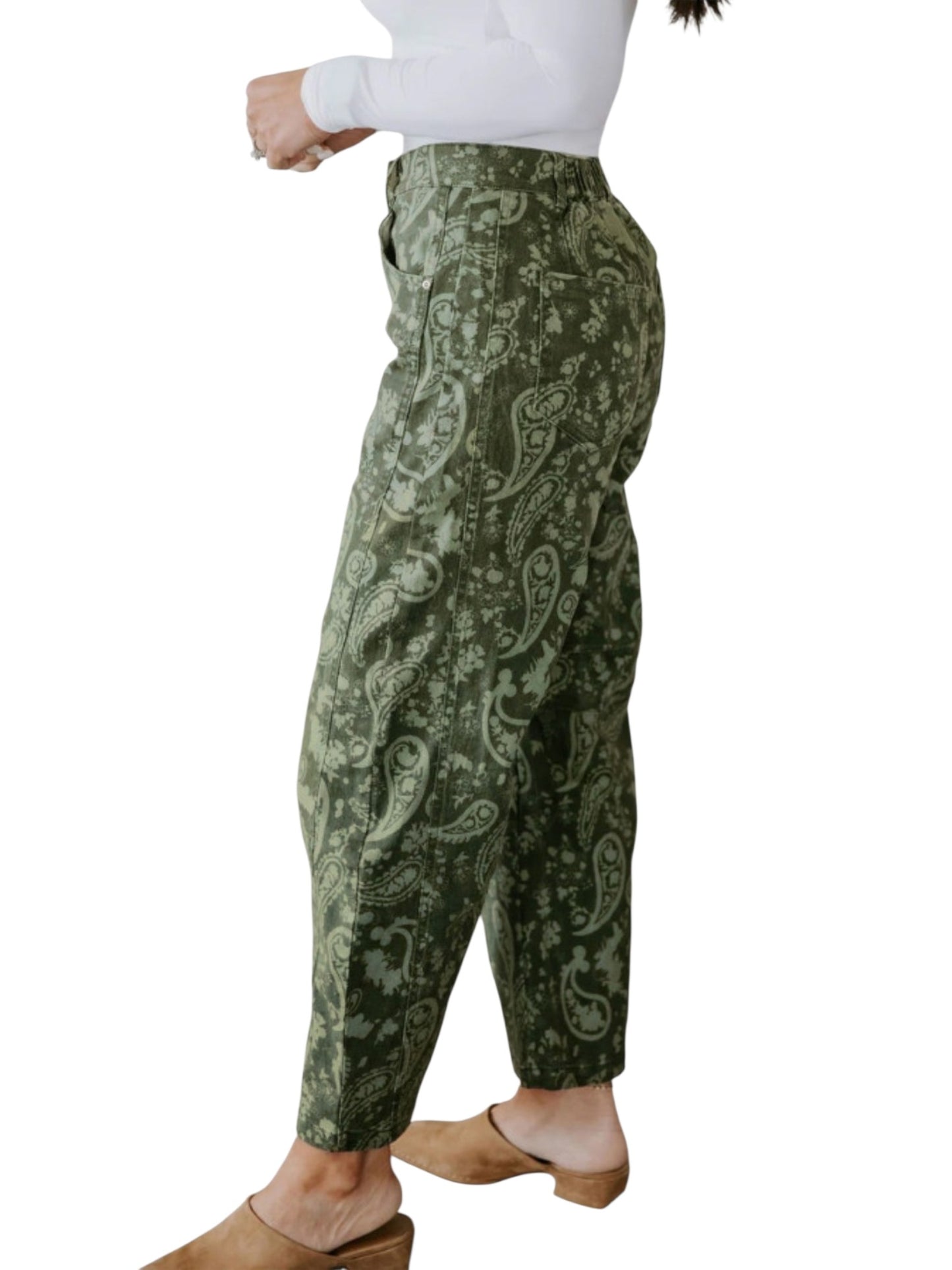 A person wearing loose, high-waisted ankle-length pants with an allover paisley pattern in olive green and light sage green, paired with a white long-sleeve top and tan mule sandals, shown against a plain white background.